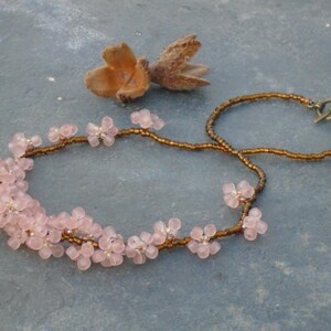 May include: A delicate pink flower necklace with a brown cord and a brass clasp. The necklace is made of small, glass beads that are shaped like flowers.