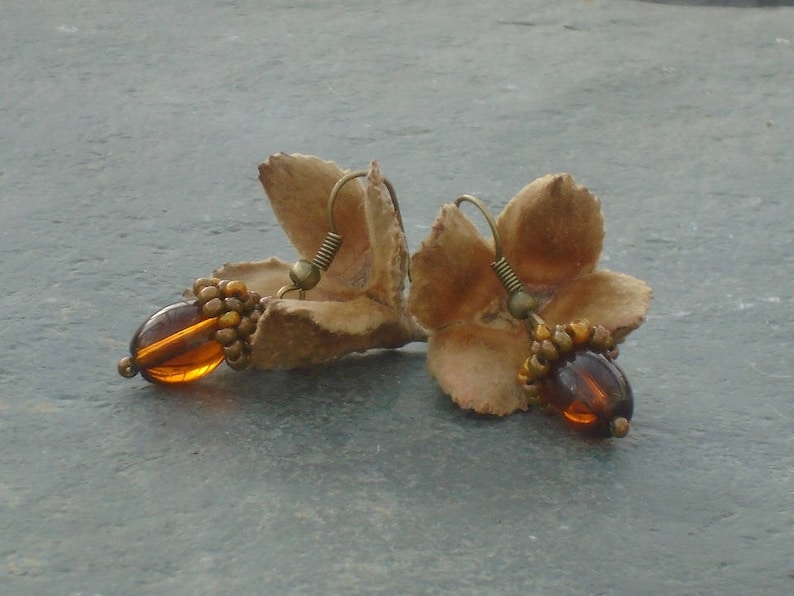 Pode incluir: Um par de brincos feitos com p&eacute;talas de flores secas e contas de vidro &acirc;mbar. Os brincos s&atilde;o de cor castanho claro e t&ecirc;m um aspeto r&uacute;stico e natural.
