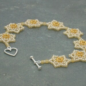 May include: A delicate bracelet made of small white and gold beads, forming a chain of flower-like shapes. The bracelet has a heart-shaped clasp.