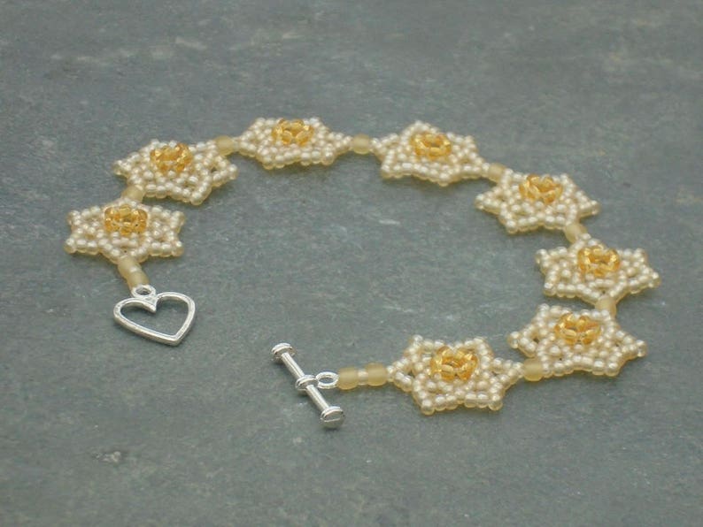 May include: A delicate bracelet made of small white and gold beads, forming a chain of flower-like shapes. The bracelet has a heart-shaped clasp.
