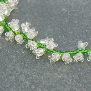 May include: A close-up of a beaded necklace with white flower-shaped beads and green beads, resembling a lily of the valley.