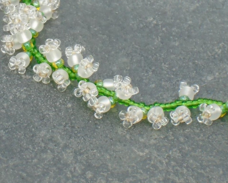 May include: A close-up of a beaded necklace with white flower-shaped beads and green beads, resembling a lily of the valley.