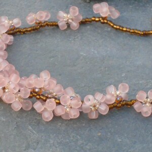 May include: A delicate necklace made with pink and brown beads. The pink beads are shaped like small flowers, and the brown beads are small and round. The necklace is strung on a brown cord.