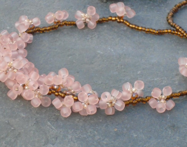 May include: A delicate necklace made with pink and brown beads. The pink beads are shaped like small flowers, and the brown beads are small and round. The necklace is strung on a brown cord.