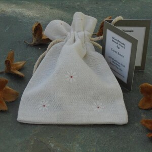 May include: A white fabric drawstring bag with small white embroidered flowers. The bag is sitting on a gray surface with brown seed pods scattered around it. A small card with the text "Handmade Bead Work by Carol Bruce Old Blakeney Road Blakeney Kent CT24 7AA" is visible in the image.