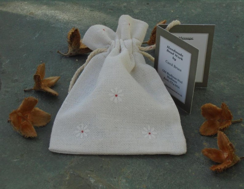 May include: A white fabric drawstring bag with small white embroidered flowers. The bag is sitting on a gray surface with brown seed pods scattered around it. A small card with the text "Handmade Bead Work by Carol Bruce Old Blakeney Road Blakeney Kent CT24 7AA" is visible in the image.