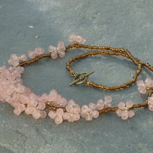May include: A delicate necklace made with pink flower-shaped beads and brown seed beads. The necklace has a simple clasp closure.