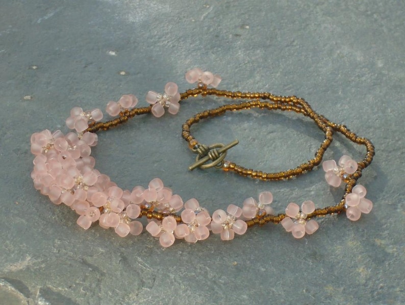 May include: A delicate necklace made with pink flower-shaped beads and brown seed beads. The necklace has a simple clasp closure.