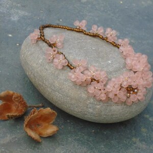 May include: A delicate pink flower necklace with brown beads. The necklace is displayed on a gray stone.