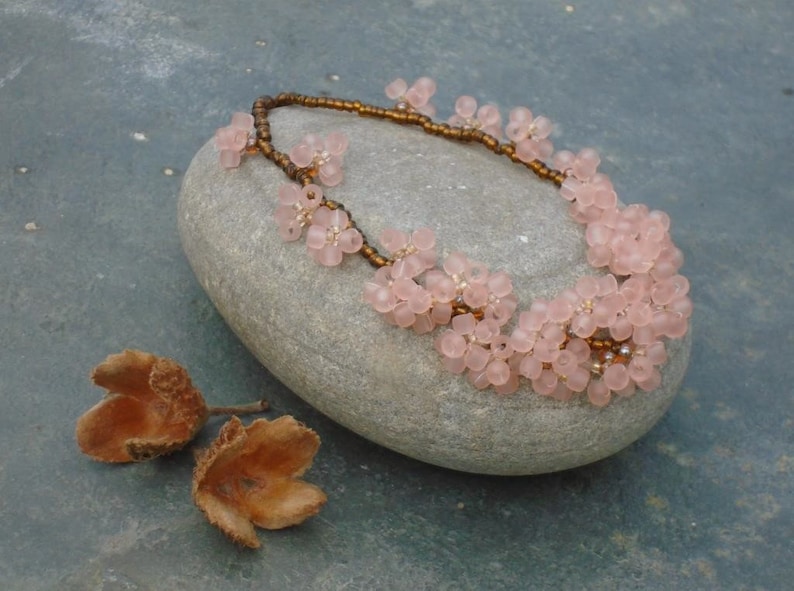 May include: A delicate pink flower necklace with brown beads. The necklace is displayed on a gray stone.