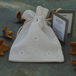 May include: A white fabric drawstring bag with small embroidered white flowers. The bag is sitting on a gray surface with brown seed pods scattered around it. A small card with the text "Handmade Bead Work by Carol Bruce Old Blakeney Road Blakeney Kent CT24 7AA" is visible in the image.
