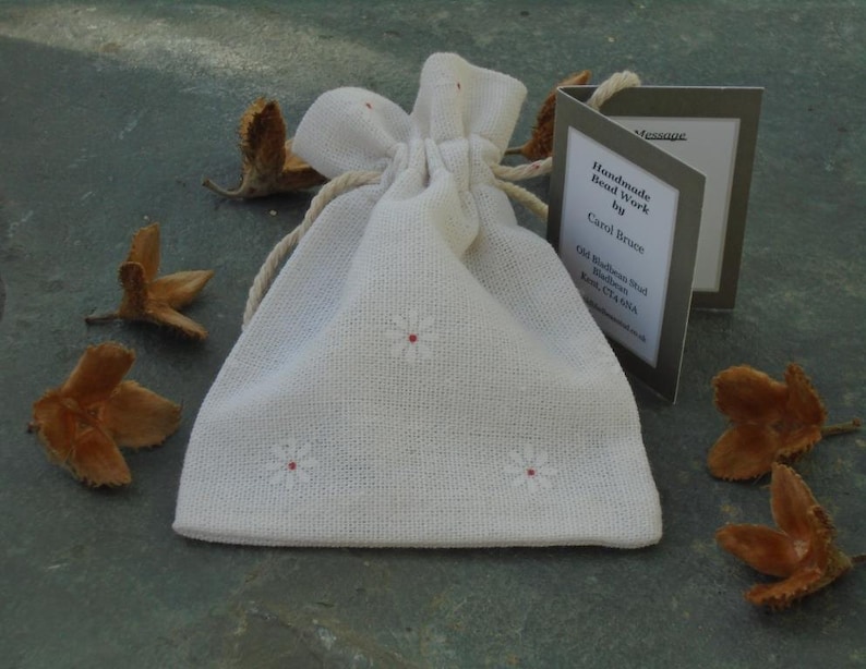 May include: A white fabric drawstring bag with small embroidered white flowers. The bag is sitting on a gray surface with brown seed pods scattered around it. A small card with the text "Handmade Bead Work by Carol Bruce Old Blakeney Road Blakeney Kent CT24 7AA" is visible in the image.