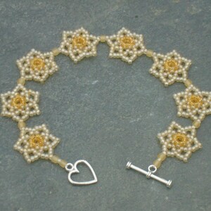 May include: A delicate bracelet made of small white and gold beads, arranged in a flower pattern. The bracelet has a heart-shaped clasp and a toggle clasp.