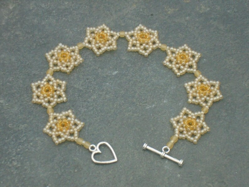May include: A delicate bracelet made of small white and gold beads, arranged in a flower pattern. The bracelet has a heart-shaped clasp and a toggle clasp.