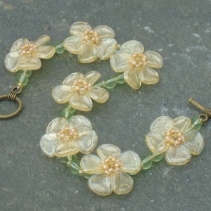 Puede incluir: Un brazalete delicado con una serie de cuentas de color amarillo pálido en forma de flor con un centro dorado, conectadas por pequeñas cuentas verdes. El brazalete tiene un cierre dorado.