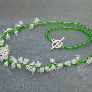 May include: A delicate necklace made with green seed beads and white glass beads that resemble lily of the valley flowers. The necklace has a silver toggle clasp.