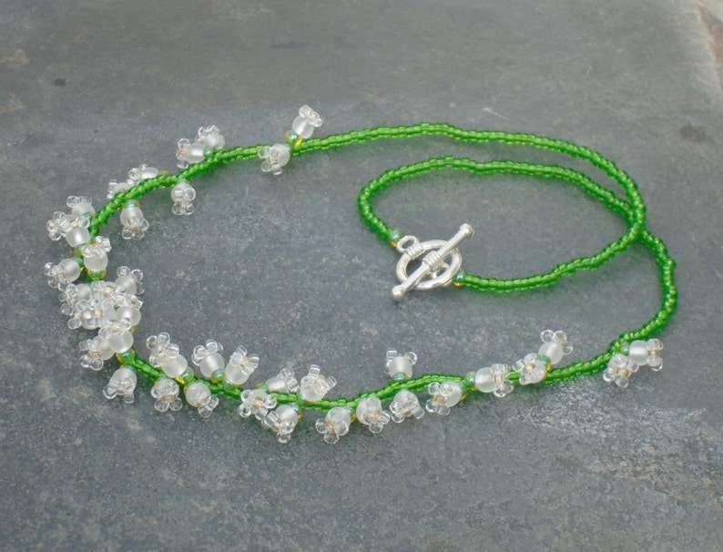 May include: A delicate necklace made with green seed beads and white glass beads that resemble lily of the valley flowers. The necklace has a silver toggle clasp.