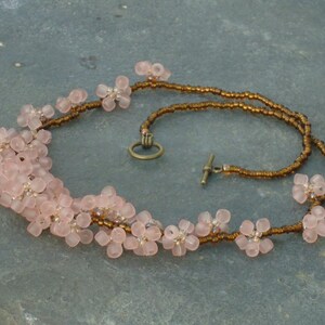 May include: A delicate pink flower necklace with brown beads. The necklace is made of small, delicate pink flowers strung together on a brown cord. The necklace has a simple clasp.