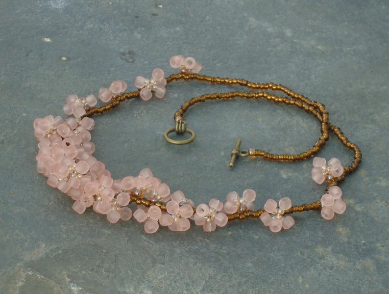 May include: A delicate pink flower necklace with brown beads. The necklace is made of small, delicate pink flowers strung together on a brown cord. The necklace has a simple clasp.