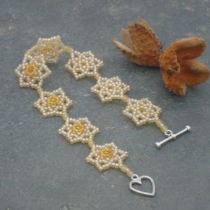 May include: A delicate beaded bracelet with a heart-shaped clasp. The bracelet is made of small white beads and features a pattern of gold-colored flowers.