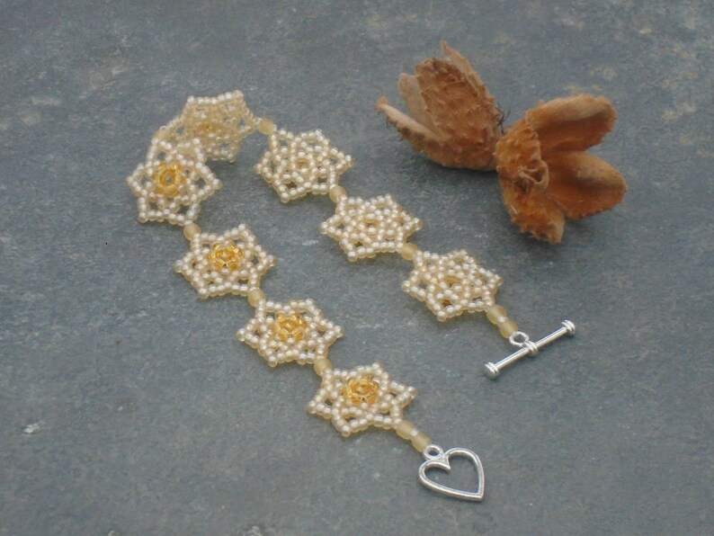 May include: A delicate beaded bracelet with a heart-shaped clasp. The bracelet is made of small white beads and features a pattern of gold-colored flowers.