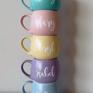 May include: Five ceramic mugs stacked on top of each other. The mugs are in different pastel colours: teal, pink, yellow, purple, and blue. Each mug has a white name written on it: Brady, Mary, Cheryl, Isabel, and Lucie.