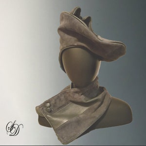 May include: A grey suede hat with a unique, folded design. The hat is worn on a mannequin with a matching grey suede scarf.
