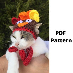 May include: A cat wearing a handmade crochet turkey hat and matching red scarf. The hat features brown, red, orange, and yellow yarn, with white eyes and an orange beak. The text "PDF Pattern" is visible.