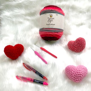 May include: A skein of pink, red, and gray yarn labeled "Sugarwheel strawberry swirl" by Yarn Bee, two pink crochet hooks, two pink highlighters, and three crocheted hearts in shades of pink and red.