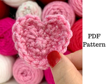 Op de afbeelding: Een roze gehaakt hart met zilverkleurige metallic draadaccenten. Het hart wordt getoond tegen een achtergrond van roze bollen garen. De tekst "PDF Pattern" is zichtbaar.
