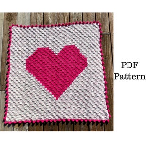 May include: A crochet blanket with a pink heart in the center. The blanket is white with a pink border. The blanket is made with a textured stitch. The text "PDF Pattern" is in the image.