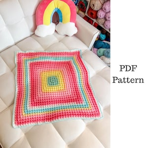 May include: A colorful, square crochet blanket with a waffle stitch pattern. The blanket features concentric squares of pink, yellow, blue, and green. A rainbow pillow and the text "PDF Pattern" are also visible.