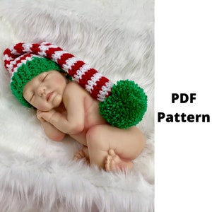 Op de afbeelding: Een pasgeboren baby draagt een rood-wit gestreepte, gehaakte elfenmuts met een groene pompon. De muts ligt op een wit, pluche oppervlak.