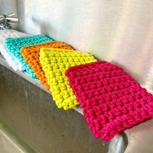 May include: Five colorful knitted dishcloths in a kitchen sink. The dishcloths are in shades of white, turquoise, orange, yellow, and pink.