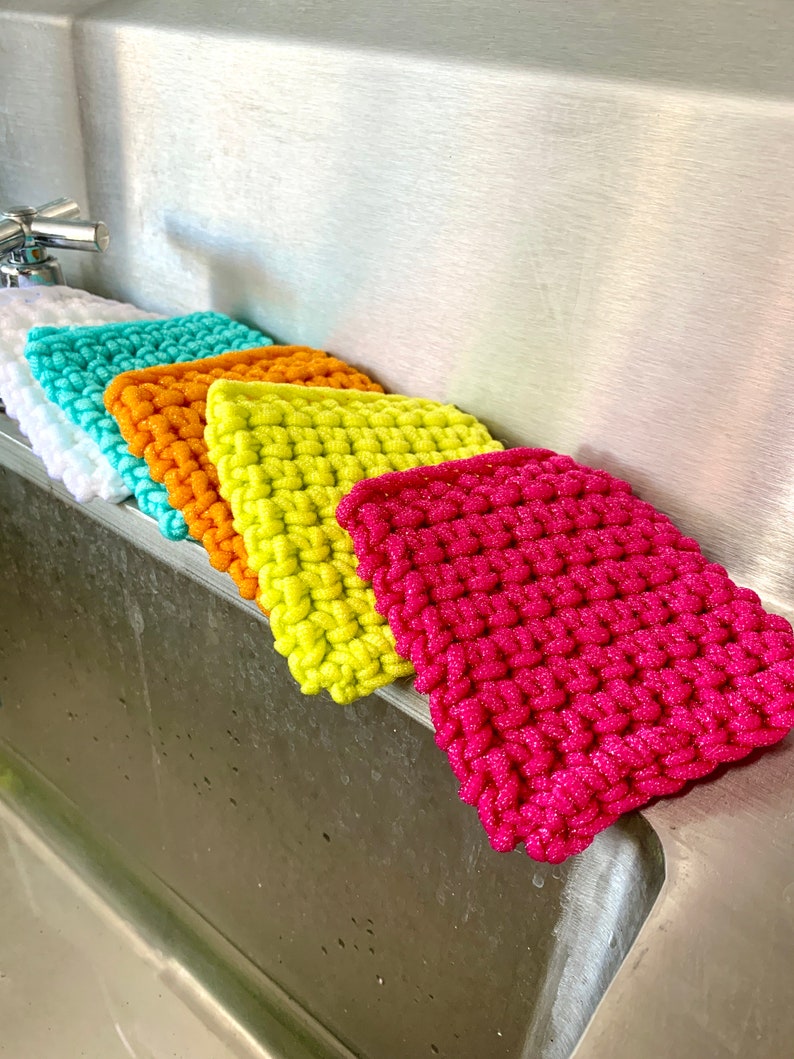 May include: Five colorful knitted dishcloths in a kitchen sink. The dishcloths are in shades of white, turquoise, orange, yellow, and pink.