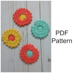 May include: Three crocheted coasters in a flower shape. One coaster is yellow with an orange centre, one is turquoise, and one is coral with a turquoise centre. The coasters are on a white wooden surface. PDF Pattern.