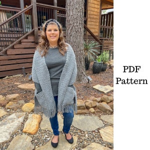 May include: A gray crochet shawl with fringe and pockets. The shawl is worn by a person with long brown hair and a black headband. The person is standing in front of a wooden building.