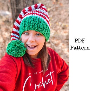 May include: A woman wearing a red sweater with the text "Tis the Season to Crochet" is wearing a green and red striped crochet elf hat with a green pom-pom.