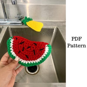 May include: A crocheted watermelon kitchen scrubber with red, white, and green yarn. The scrubber is in a stainless steel kitchen sink.