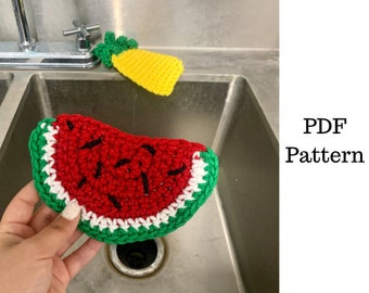 Puede incluir: Una esponja de cocina de ganchillo en forma de sandía con hilo rojo, blanco y verde. La esponja está en un fregadero de cocina de acero inoxidable.