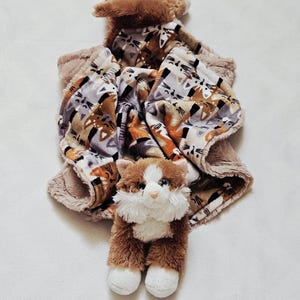 May include: A plush cat toy with a brown and white body and a soft, patterned blanket. The blanket features a repeating cat face design in shades of brown, gray, and orange. The toy is designed for babies and young children.
