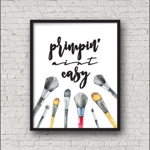 May include: A black framed print with the text "primpin' ain't easy" in black cursive font. The print features a watercolor illustration of seven makeup brushes in various sizes and colors.