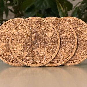 May include: Four round cork coasters with a detailed map of Atlanta, GA etched into the surface. The city's name is printed along the bottom edge of each coaster. The coasters are a natural cork color and are arranged in a row.