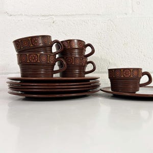 Könnte beinhalten: Ein Satz brauner Keramikbecher und Untertassen. Die Tassen und Untertassen haben ein dekoratives Blumenmuster. Mehrere Tassen sind gestapelt, mit einer separaten Tasse und Untertasse. Das Set steht auf einer weißen Oberfläche.