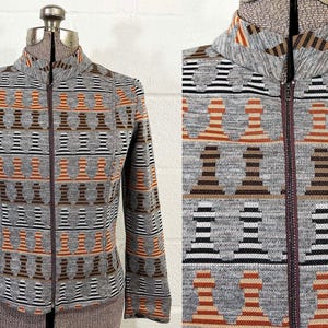May include: A gray zip-up jacket with a repeating pattern of chess piece-like shapes in brown, orange, black, and white. The jacket has a high collar and a dark brown zipper. The fabric appears to be a knit material.