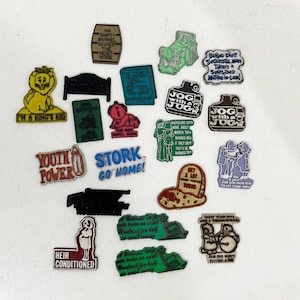 May include: A collection of twenty-one assorted novelty pins featuring various shapes, colors, and humorous text. The pins include designs like a yellow lion, a green lawnmower, a brown jug, and text-based slogans such as Youth Power and Stork Go Home.