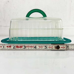 May include: A clear plastic butter dish with a teal green lid and base. The lid has a handle and the base has a beaded edge.