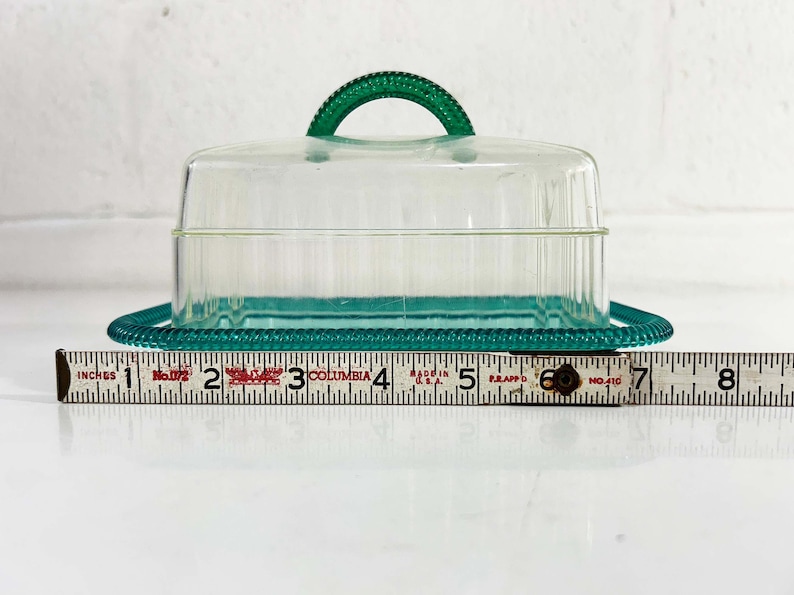 May include: A clear plastic butter dish with a teal green lid and base. The lid has a handle and the base has a beaded edge.