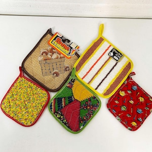 Könnte beinhalten: Fünf verschiedene Topflappen. Einer ist braun mit einem gesteppten Muster und Pilzen in einem Korb. Ein anderer ist gelb mit einem Blumenmuster. Ein dritter ist grün mit einem Patchwork-Muster. Ein vierter ist weiß mit braunen Streifen. Der letzte ist rot mit einem Muster aus Teekannen. Der braune Topflappen hat ein Etikett mit der Aufschrift "Flame Resistant".