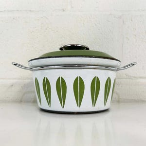 Puede incluir: Una olla de esmalte blanco con tapa verde y diseño de hojas verdes. La olla tiene un borde plateado y dos asas plateadas.